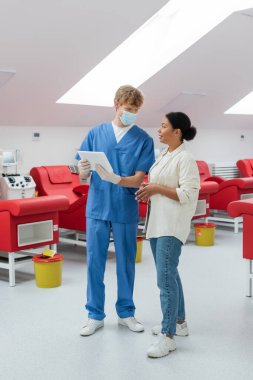 Mavi üniformalı kızıl saçlı doktor, tıbbi maske ve lateks eldivenler elinde dijital tablet ve tıbbi sandalyelerin yanında çok ırklı bir kadınla konuşmak, kan nakli makinesi ve laboratuardaki çöp kovaları.
