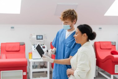 Tıbbi maskeli kızıl saçlı doktor, lateks eldivenler ve mavi üniformalı. Klinikteki bulanık tıbbi sandalyelerin ve otomatik kan nakli makinesinin yanındaki çok ırklı kadınlara boş ekranlı dijital tablet gösteriyor.