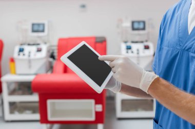 partial view of doctor in latex gloves pointing at digital tablet with blank screen near blurred medical chair and blood transfusion machine on blurred background in clinic clipart