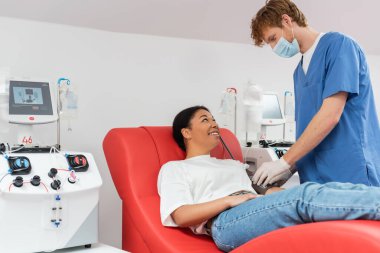 Mavi üniformalı kızıl saçlı doktor, tıbbi maske ve lateks eldivenler. Laboratuvardaki otomatik kan nakli makinelerinin yanında tıbbi sandalyede gülümseyen çok ırklı kadının kolunda kan basıncı kelepçesi var.