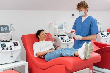Tıbbi maskeli kızıl saçlı doktor, mavi üniformalı ve lateks eldivenli. Kan bağışı merkezindeki otomatik nakil makinelerinin yanındaki tıbbi sandalyede çok ırklı kadının yanında dijital tablet kullanıyorlar.
