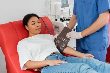 Mavi üniformalı ve lateks eldivenli bir sağlık görevlisi hastanedeki otomatik nakil makinesinin yanında oturan çok ırklı bir kadının koluna kan basıncı kelepçesi takıyor. 