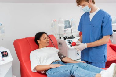 Mavi üniformalı doktor, tıbbi maske ve lateks eldivenli doktor kan basıncı kelepçeli tıbbi kauçuk kürenin yanında kan nakli makinesinin yanındaki ergonomik sandalyede oturuyor.
