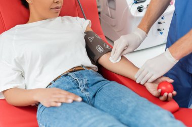 Doktorun lateks eldivenli, elinde alkol pedi olan ve çok ırklı bir kadının kolunu silen, plastik topla rahat tıbbi koltukta oturan, kan bağışı merkezinde ergonomik tasarımı olan bir doktor görüntüsü.