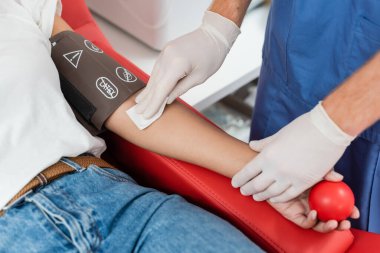 Kauçuk eldivenli doktorun elinde alkol pedi ve kan bağışı merkezinde tıbbi sandalyede otururken elinde lastik top tutan çok ırklı kadının kollarını silmesi. 