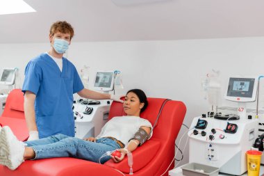 Tıbbi maskeli bir doktor, mavi üniformalı ve lateks eldivenli. Kan nakli makinelerinin yanındaki kameraya bakıyor. Klinikte kan bağışlarken çok ırklı bir kadın koltuğunda oturuyor.
