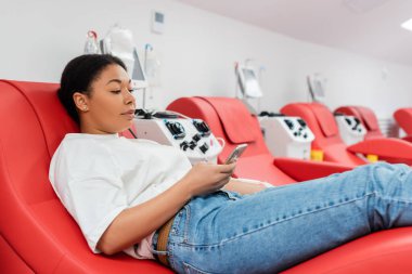 Hastanede kan bağışı sırasında ergonomik tıbbi sandalyenin yanında otururken cep telefonuyla mesajlaşan genç çok ırklı kadın, tıbbi prosedür...
