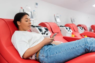 young multiracial woman messaging on mobile phone while sitting on ergonomic medical chair near transfusion machine during blood donation in hospital, medical procedure clipart