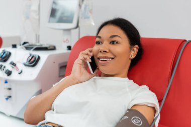 young and cheerful multiracial woman talking on smartphone while sitting on medical chair near blurred transfusion machine in blood donation center  clipart