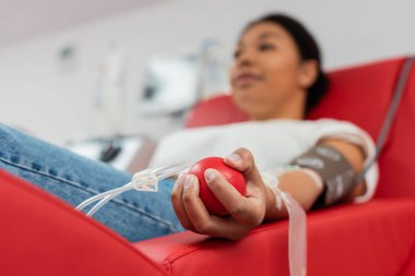 Tıbbi kauçuk topunun seçici odağı, laboratuarda rahat bir tıbbi sandalyede otururken kan bağışı yapan çok ırklı bir kadının elinde bulanık bir arka plan.