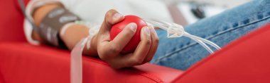 partial view of multiracial woman with transfusion set holding rubber ball while sitting on ergonomic medical chair during blood donation in laboratory, banner clipart