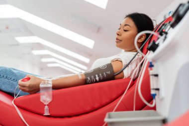 side view of multiracial woman in pressure cuff and transfusion set holding rubber ball while sitting on comfortable medical chair near blurred transfusion machine in clinic clipart