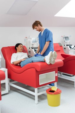 Mavi üniformalı kızıl saçlı doktor ve otomatik kan nakli makinelerinin yanında lateks eldivenler ve hastane hastanesinde kan bağışı yapan çok ırklı kadın.