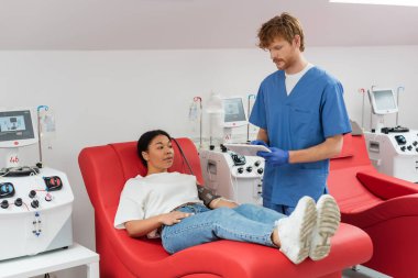 Mavi üniformalı, lateks eldivenli, transfüzyon makinesinin yanında dijital tablet kullanan kızıl saçlı doktor ve klinikteki tıp koltuğunda oturan çok ırklı kadın.
