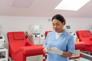 Mavi üniformalı çok ırklı hemşire ve lateks eldivenli hemşire kan bağışı merkezinde tıbbi koltukların yanında cep telefonuyla ve kan nakli makineleriyle mesajlaşıyor.