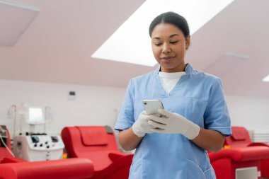 Mavi üniformalı ve lateks eldivenli pozitif çok ırklı doktor kan bağışı merkezindeki bulanık arka planda kan nakli makinesi ve tıbbi sandalyelerin yanında akıllı telefondan mesajlaşıyor.