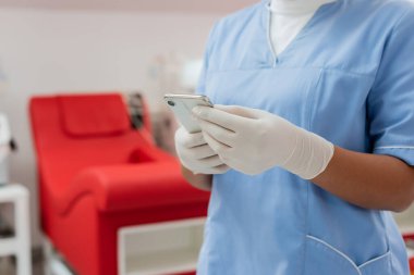 Mavi üniformalı ve steril lateks eldivenli hemşirenin kısmi görüntüsü. Kan nakli merkezindeki bulanık tıbbi koltuğun yanında cep telefonuyla mesajlaşıyor. 