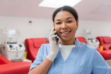 happy multiracial healthcare worker in blue uniform and latex glove talking on mobile phone near medical chairs and transfusion machines in blood donation center clipart