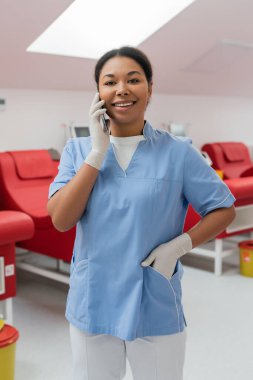 Mavi üniforma cebinde duran lateks eldivenli neşeli çok kültürlü hemşire laboratuvarda bulanık tıbbi sandalyelerin yanında akıllı telefonla konuşuyor.