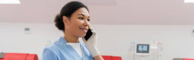happy multiracial healthcare worker in blue uniform smiling during conversation on mobile phone in blood donation center on blurred background, banner clipart