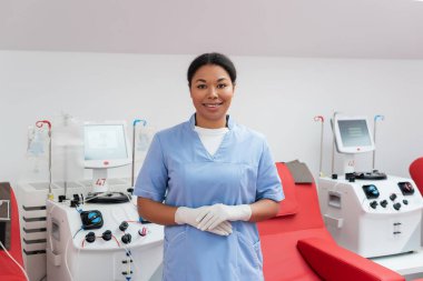 Mavi üniformalı, lateks eldivenli neşeli hemşire kan bağışı merkezindeki otomatik nakil makinelerinin ve ergonomik tıbbi sandalyelerin yanındaki kameraya bakıyor.