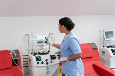 Mavi üniformalı ve lateks eldivenli genç çok ırklı doktor kan bağışı merkezinde tıbbi sandalyelerin yanında dokunmatik ekranlı transfüzyon makinesi kullanıyor.