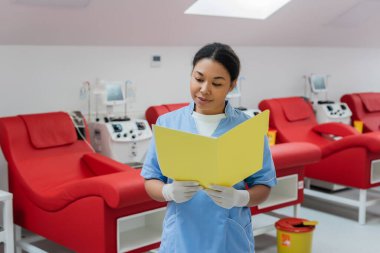 multiracial healthcare worker in blue uniform and latex gloves reading medical charts next to blurred transfusion machines, medical chairs and trash bucket in laboratory clipart