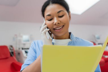Mavi üniformalı, lateks eldivenli neşeli hemşire akıllı telefondan konuşuyor ve kan bağışı merkezinde bulanık kan nakli makineleri ve tıbbi sandalyelerin yanındaki kağıt klasöre bakıyor.
