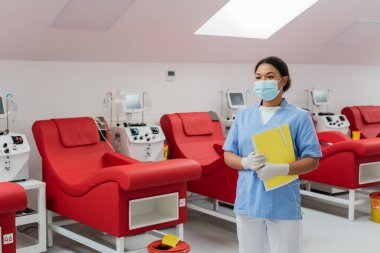 Üniformalı çok ırklı sağlık çalışanı, tıbbi maske ve lateks eldivenler elinde kağıt klasör ve kalemle ergonomik tıbbi sandalyelerin yanında duruyor ve kan bağışı merkezinde nakil makineleri var.