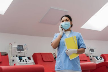Mavi üniformalı çok ırklı hemşirenin düşük açılı görüntüsü, tıbbi maske ve lateks eldivenler akıllı telefondan konuşuyor, elinde kağıt klasör ve hastane koltuklarının yanında kalem ve kan nakli makineleri var.