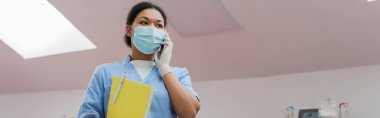 Mavi üniformalı çok ırklı hemşirenin düşük açılı görüntüsü, tıbbi maske ve lateks eldiven elinde kağıt klasör ve cep telefonu ile kan bağışı merkezinde konuşma, afiş.