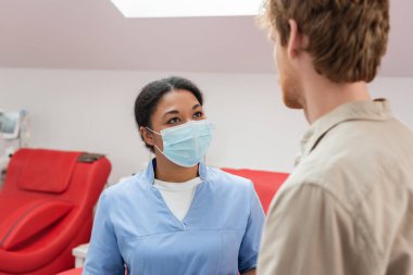 Mavi üniformalı ve tıbbi maskeli çok ırklı bir sağlık çalışanı kan bağışı merkezinde tıbbi sandalyelerin yanında bulanık ön planda genç kızıl saçlı gönüllülerle konuşuyor.