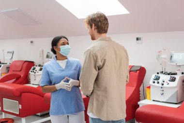 Üniformalı, tıbbi maskeli ve lateks eldivenli çok ırklı sağlık görevlisi klinikteki tıbbi sandalyelerin yanında kızıl saçlı adamla ve otomatik kan nakli makineleriyle konuşuyor.