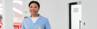 joyful multiracial healthcare worker in blue uniform looking at camera while standing in waiting area of modern contemporary blood donation center, banner, door on background  clipart
