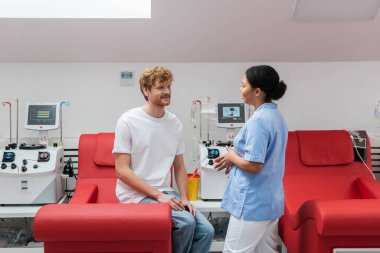 multiracial nurse in uniform talking to positive redhead volunteer sitting on medical chair near transfusion machines and plastic cup in blood donation center clipart