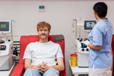 Mavi üniformalı çok kültürlü hemşire plastik bardağın yanında kan nakli makinesi çalıştırıyor ve kızıl saçlı gönüllü tıbbi sandalyede oturuyor ve kan bağışı merkezindeki kameraya bakıyor.