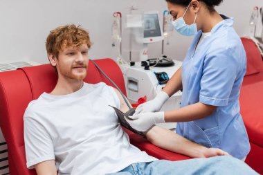 Tıbbi maskeli ve lateks eldivenli çok ırklı hemşire klinikteki nakil makinesinin yanında oturan kızıl saçlı donörün kolunda kan basıncı kelepçesi ayarlıyor.