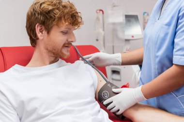 Mavi üniformalı ve lateks eldivenli çok ırklı hemşire laboratuardaki kan nakli makinesinin yanında kızıl kan donörünün koluna kan basıncı kelepçesi takıyor.