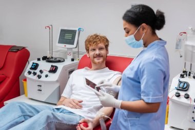 multiracial nurse in blue uniform and medical mask standing with digital tablet near smiling redhead volunteer on comfortable chair next to automated equipment in blood donation center clipart