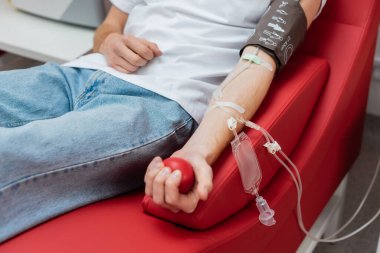 Kan nakli seti olan genç bir adamın kısmi görüşü ergonomik ve rahat koltukta oturan kan basıncı kelepçesi ve modern tıp laboratuarında lastik top sıkması.