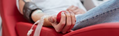partial view of young volunteer sitting on comfortable ergonomic chair and squeezing rubber ball during medical procedure in blood donation center, banner clipart