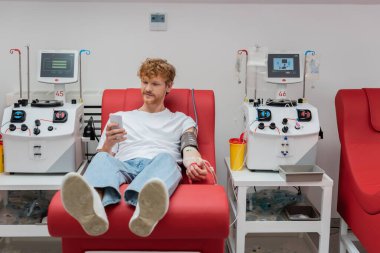 Genç kızıl saçlı gönüllü ergonomik tıbbi sandalyede oturuyor ve klinikteki otomatik ekipman ve plastik bardağın yanındaki kan bağışı sırasında akıllı telefondan mesajlaşıyor.