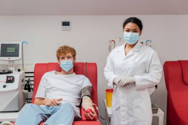 Tıbbi maskeli çok ırklı doktor, kan basıncı kelepçesi ve kan nakli setiyle gönüllü olarak laboratuardaki otomatik ekipman ve plastik bardağın yanında oturuyor.