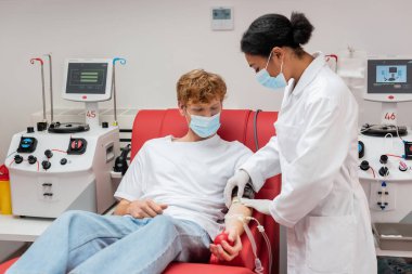 Sağlık maskeli kızıl saçlı gönüllü otomatik ekipmanların yanında rahat koltukta oturuyor ve beyaz önlüklü çok ırklı doktor kan bağışı merkezinde kan naklini ayarlıyor.