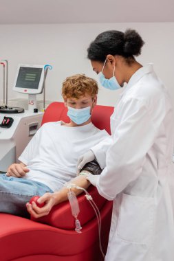 Beyaz üniformalı ve tıbbi maskeli çok ırklı doktor kan bağışı merkezindeki tıbbi ekipmanların yanında oturan kan nakli seti olan kızıl saçlı adamın koluna yara bandı yapıştırıyor.