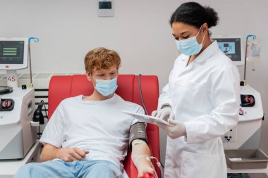 Beyaz önlüklü çok ırklı doktor, tıbbi maske ve lateks eldivenler, klinikte kan nakli makinelerinin yanında oturan kan basıncı kelepçeli kızıl saçlı donörün yanında dijital tablet kullanıyor.