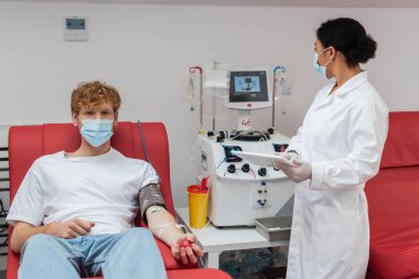 Tıbbi maskeli ve kan basıncı kelepçeli kızıl saçlı donör dijital tabletli, kan nakli makineli ve laboratuardaki plastik bardaklı çok ırklı doktorun yanındaki tıbbi sandalyede kameraya bakıyor.