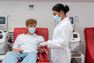 Tıbbi maskeli ve beyaz önlüklü çok ırklı doktor kan bağışı merkezinde kan nakli makinelerinin yanındaki ergonomik sandalyede oturan kızıl saçlı gönüllüye kauçuk top veriyor.