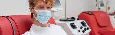Tıbbi maskeli genç ve kızıl saçlı gönüllü, pozitif bakışlı, çağdaş nakil makinesinin yanında kan bağışı yapan ve laboratuardaki kameraya bakan, afiş