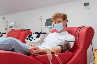 Tıbbi maskeli genç kızıl saçlı donör ve kan nakli seti klinikteki kan bağışı prosedürü sırasında otomatik ekipmanların yanında rahat koltukta otururken lastik topu sıkıyor.